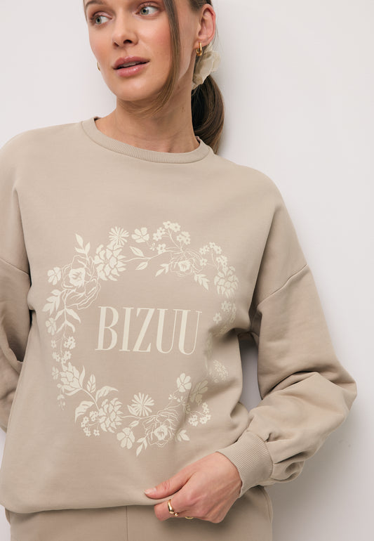 Zbliżenie na modelkę w beżowej bluzie ORISA z ozdobnym nadrukiem w kształcie wieńca.