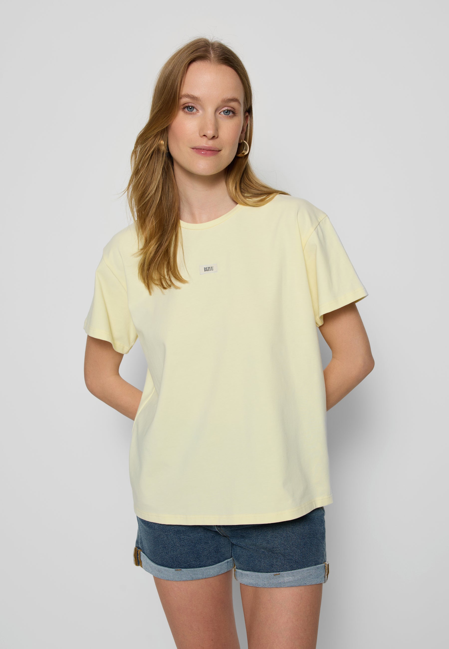 Modelka na białym tle z rękoma z tyłu w masełkowym T-shircie TISHALLE i jeansowych szortach MENORCA.