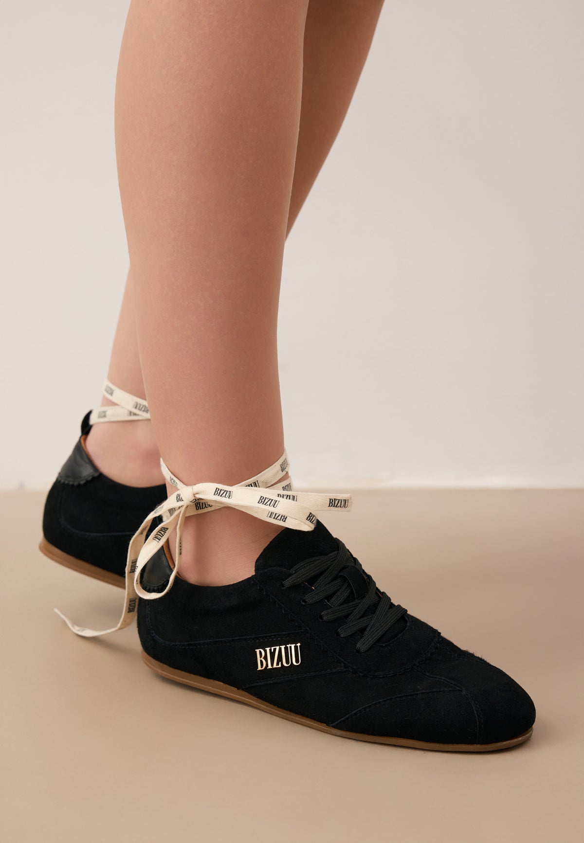 Zbliżenie na nogi modelki stojącej pod kątem w czarnych zamszowych sneakersach LEOLA ze złotym logo z boku i przewiązaną nad kostką taśmą z logo Bizuu.