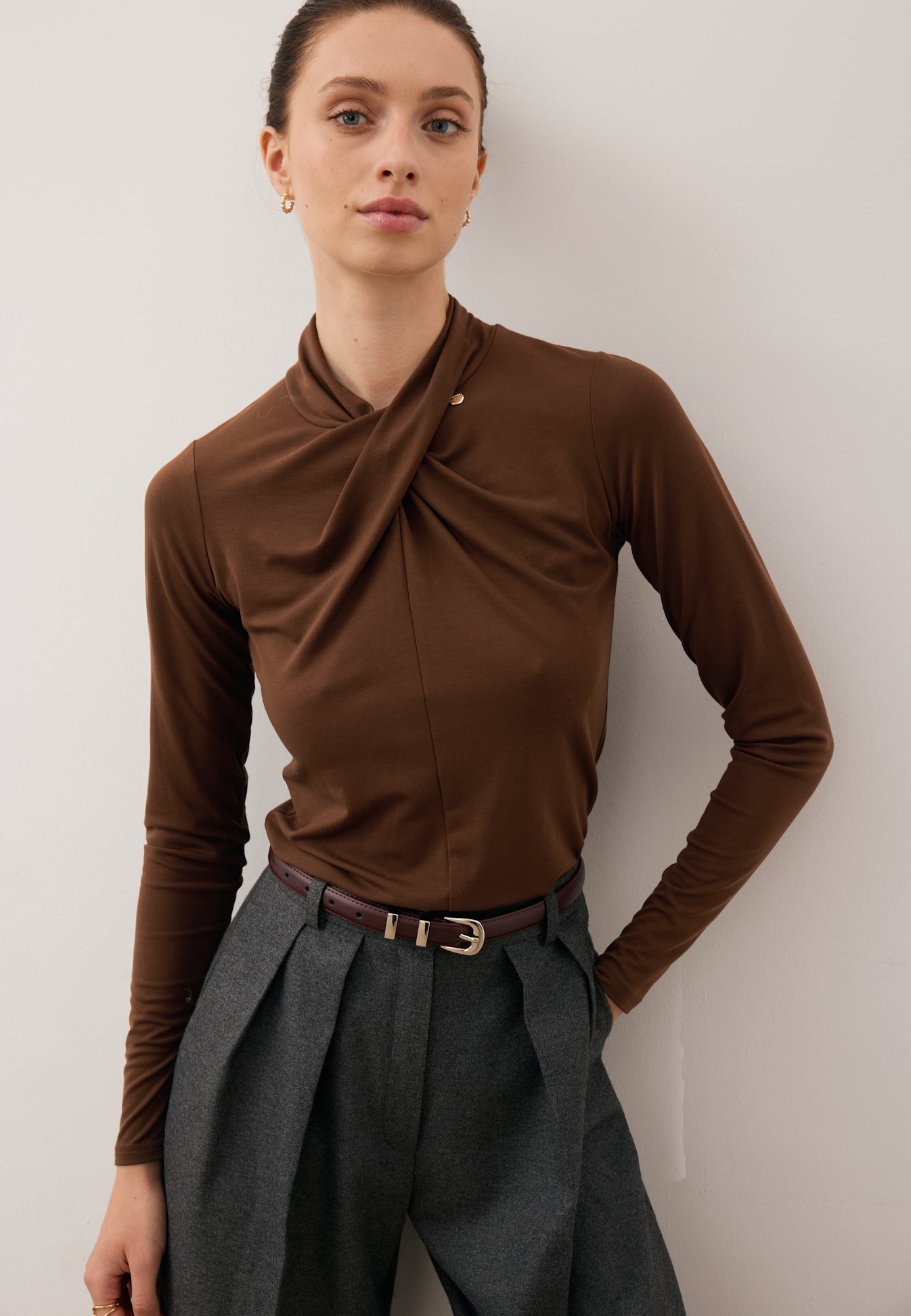 Zbliżenie na modelkę z jedną ręką z tyłu ubraną w czekoladowy longsleeve z przepleceniem PINOWA oraz szerokie spodnie BANOS.