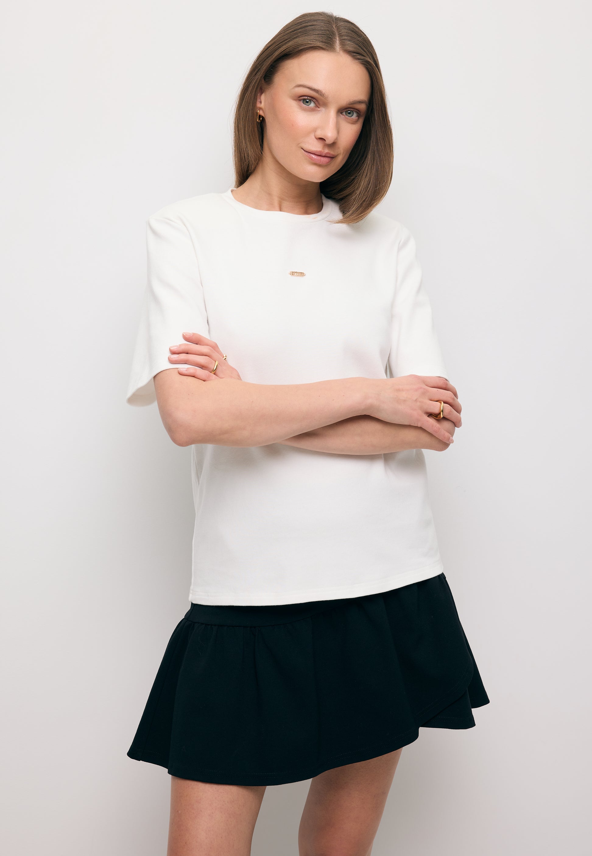 Modelka ze skrzyżowanymi rękoma ubrana w biały T-shirt z ozdobnymi poduszkami MELISA oraz czarną mini spódniczkę TULIAN.