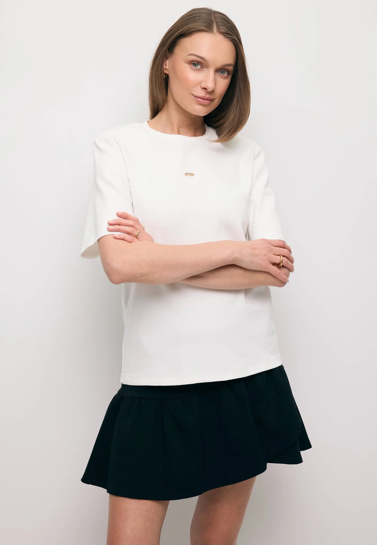 Modelka ze skrzyżowanymi rękoma ubrana w biały T-shirt z ozdobnymi poduszkami MELISA oraz czarną mini spódniczkę TULIAN.