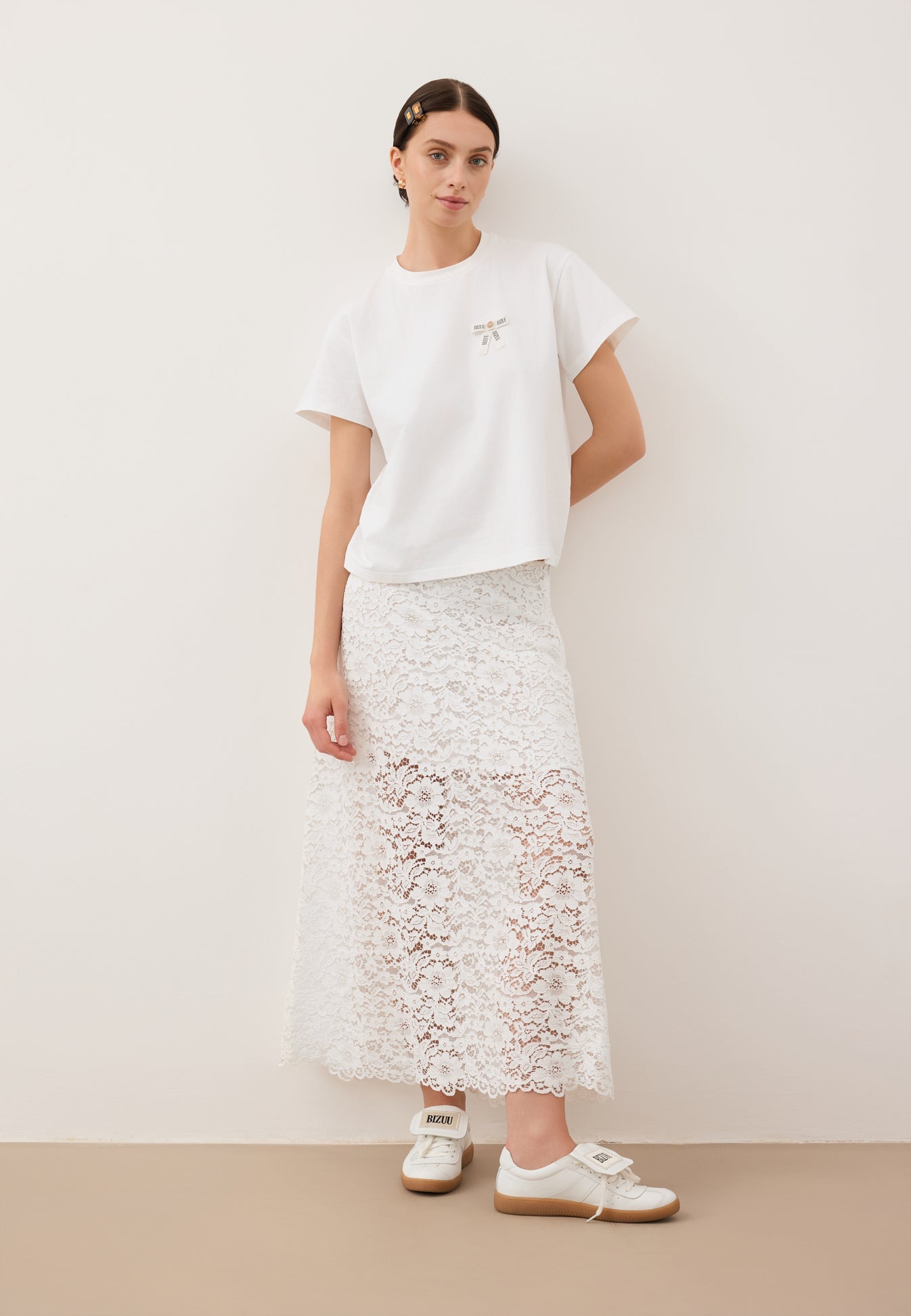 Modelka oparta o ścianę z jedną ręką zgiętą do tyłu ubrana w T-shirt z logowaną kokardką TISHALLE, białą, koronkową spódniczkę MILKY i buty MOBBY.
