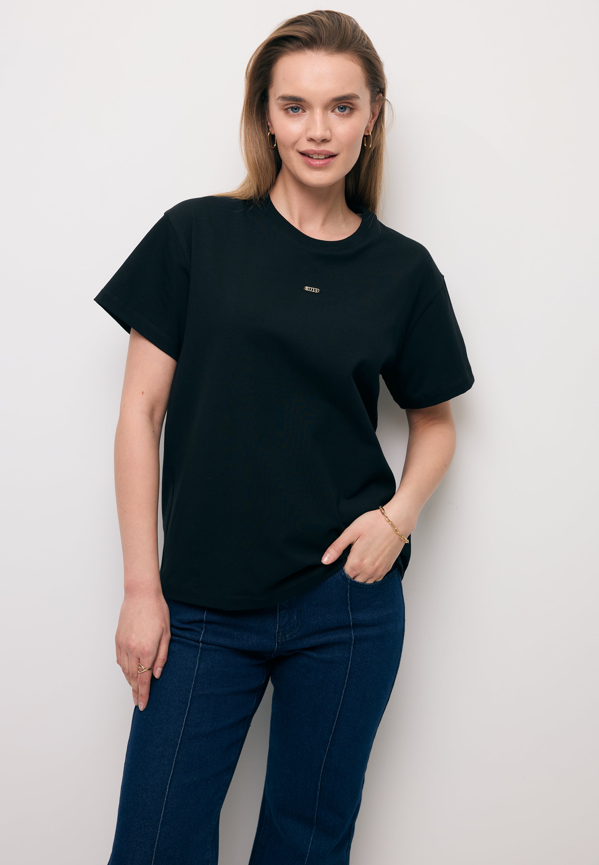 Modelka na tle białej ściany z jedną ręką na biodrze. Ubrana w czarny T-shirt TISHAL i koronkową spódniczkę FLACE.